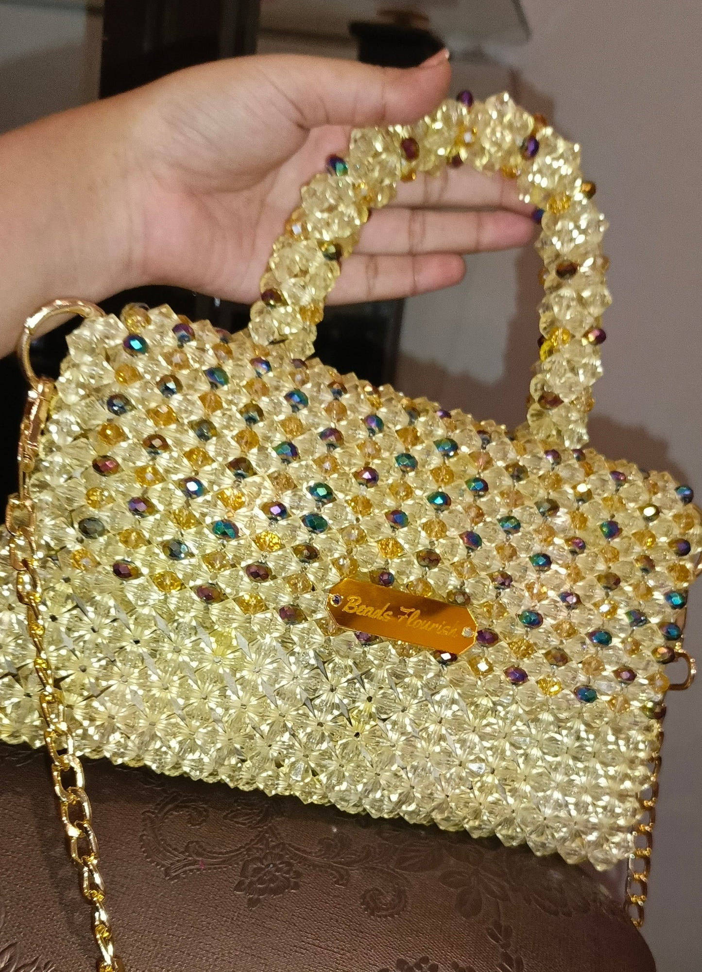 Gold beaded handbag with a chain strap held by a hand on a brown textured surface.